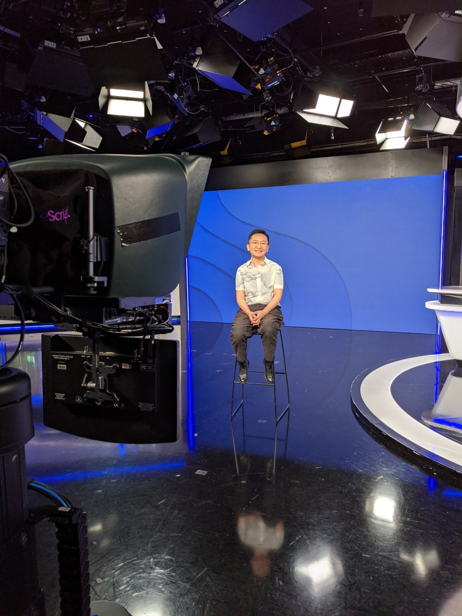 Jinghua in a white shirt and black pants, sitting on a high stool on a tv set with a blue background and a camera in the foreground.