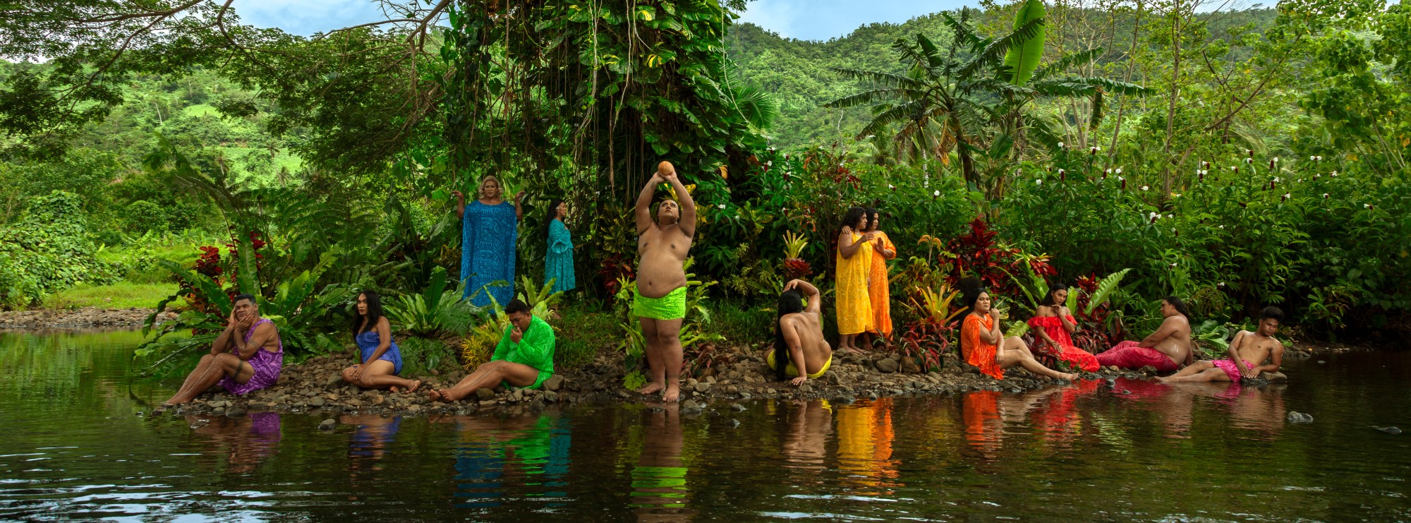 Tableau photograph of people posed on a river bank, dressed in colours of the rainbow.