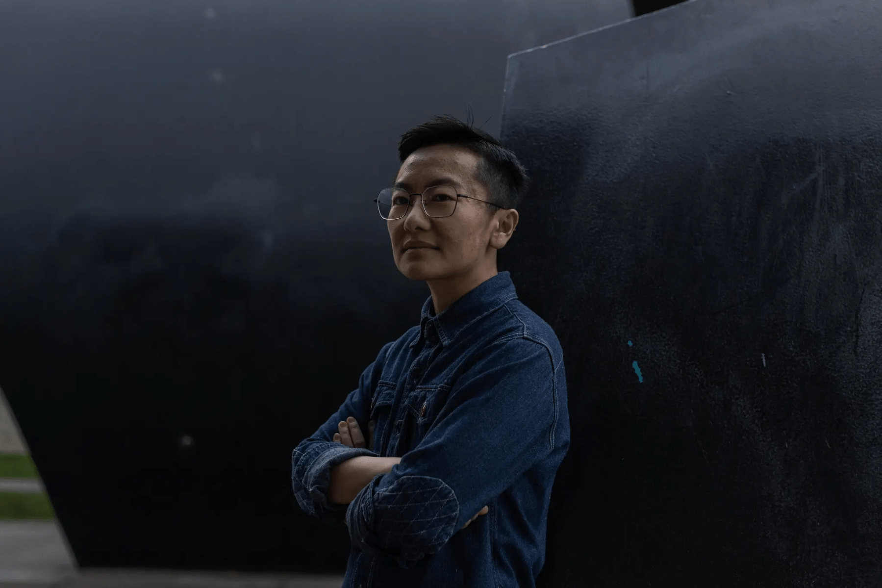 3/4 view of an Asian person with short black hair and wire glasses, wearing a dark denim shirt, standing with arms crossed against a black sculpture